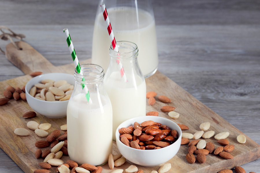 Recept voor alcoholvrije amandelcocktail Sfeerbeeld "alcoholvrije amandelcocktail": twee gevulde flessen met brede hals en kleurrijke rietjes staan op een houten plank met amandelen