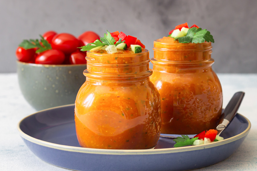 Sfeerbeeld: Twee potten met schroefdeksel gevuld met rode gazpacho op een blauw bord, op de achtergrond een schaal met tomaten
